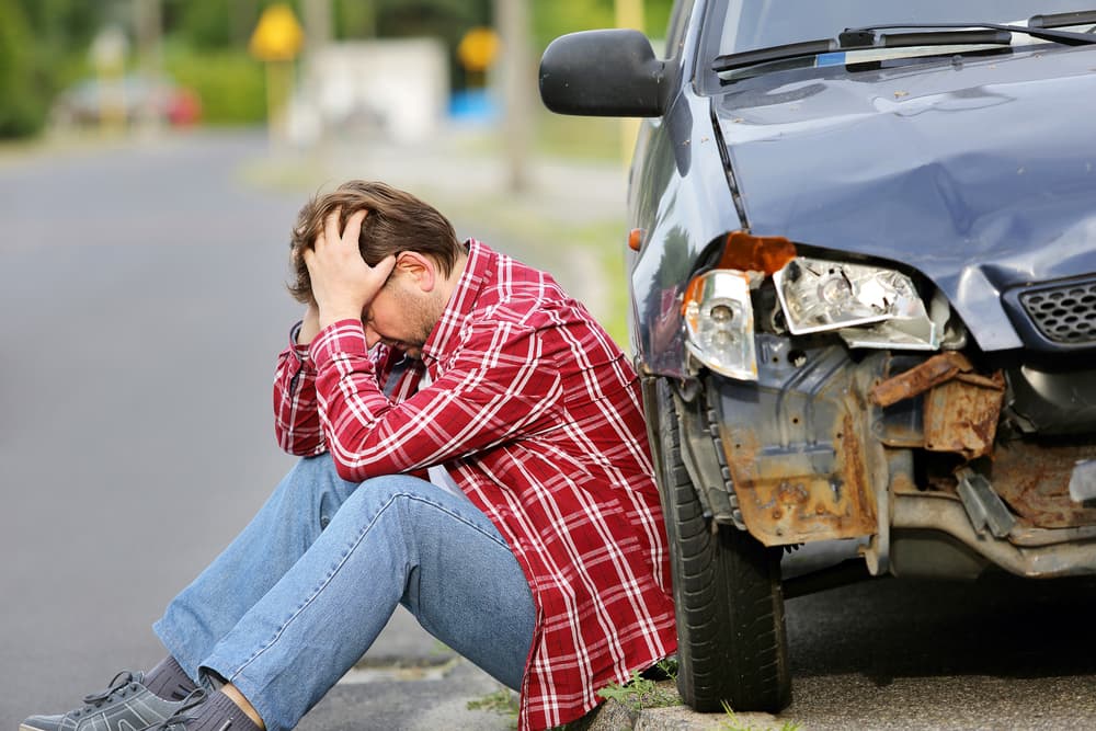 A man sits next to his damaged car following an accident, appearing distressed and reflecting on the incident.