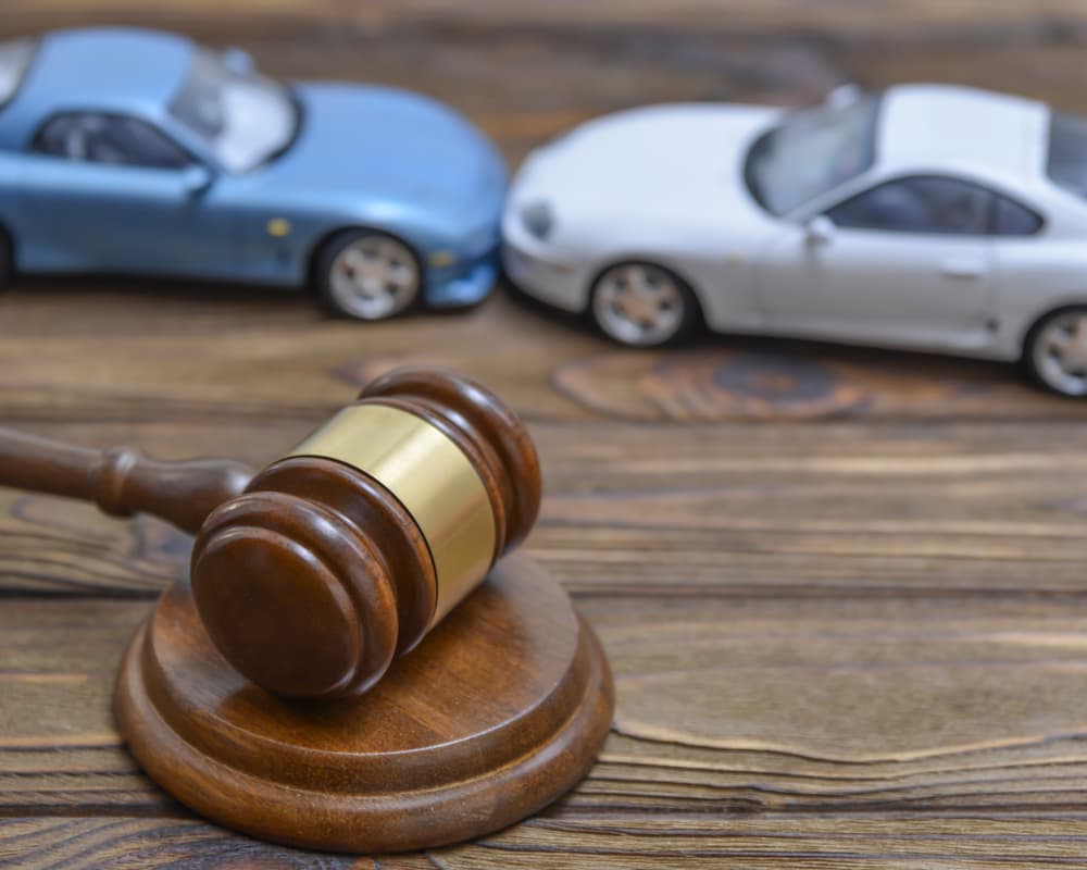 A judge's gavel next to two collided cars placed on a wooden surface.