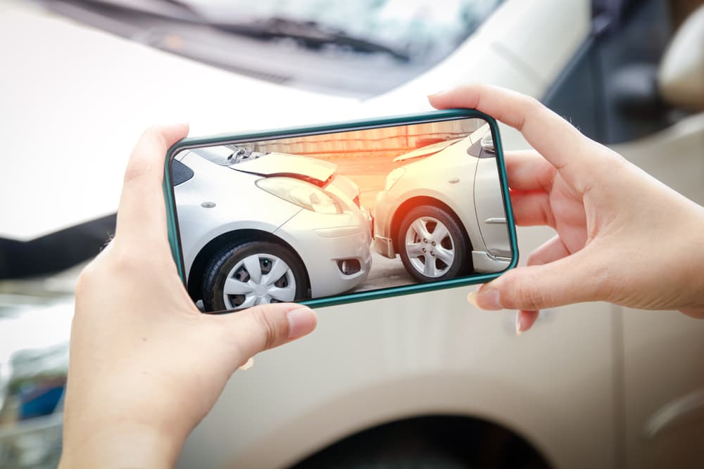 A woman uses her smartphone to take photos of a car accident scene, gathering evidence for a car insurance claim.