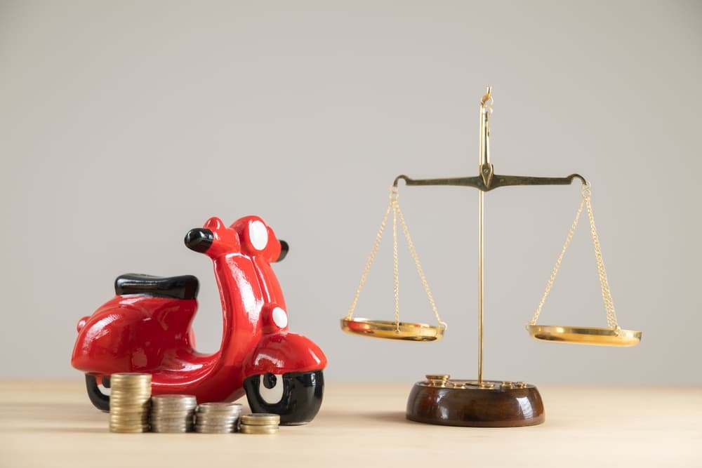 A red scooter beside a balance scale symbolizes financial and legal considerations in motorcycle accidents.